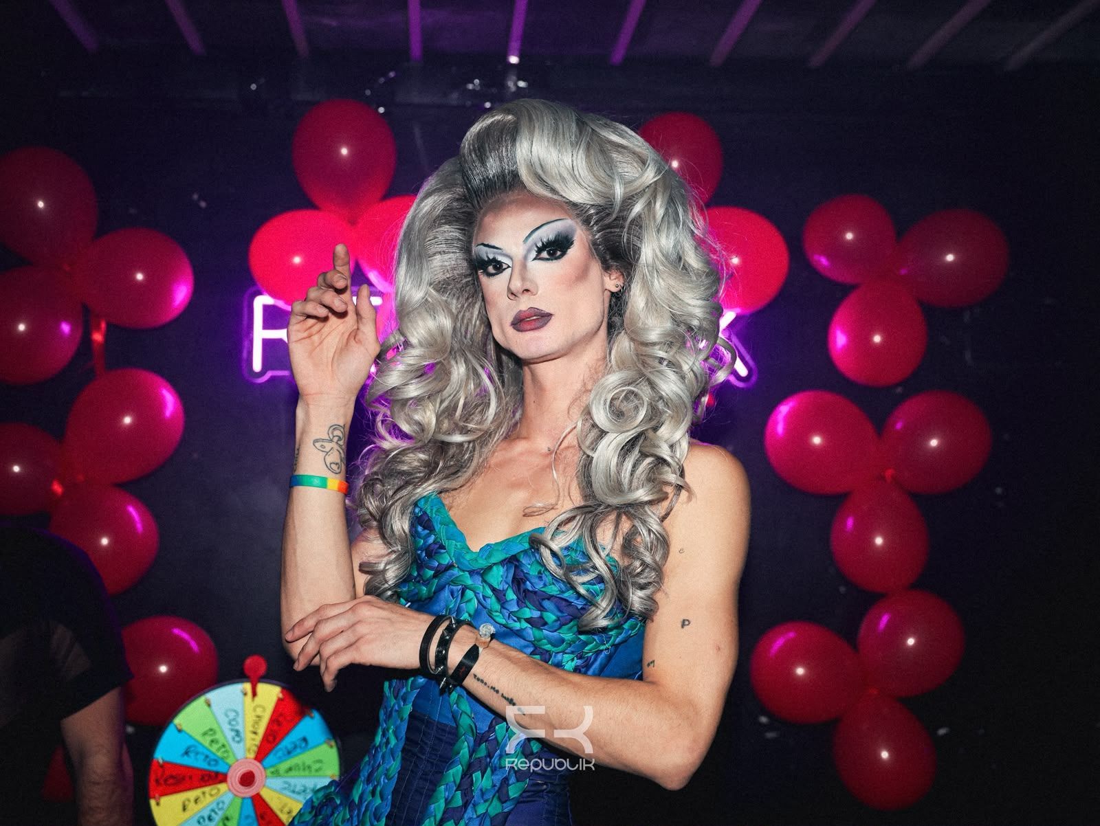 Persona con peluca plateada y vestido verde azulado en una fiesta, sosteniendo un accesorio rosa con globos al fondo