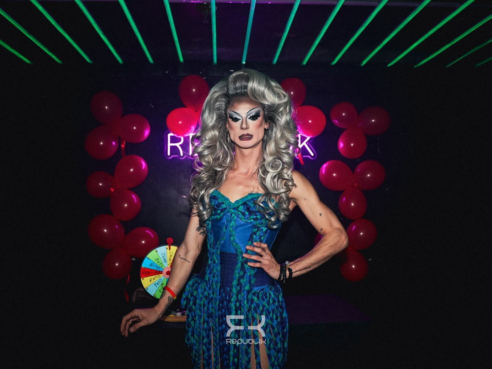 Una persona con un vestido azul y una peluca plateada posa frente a un fondo de globos iluminado con luces de neón.