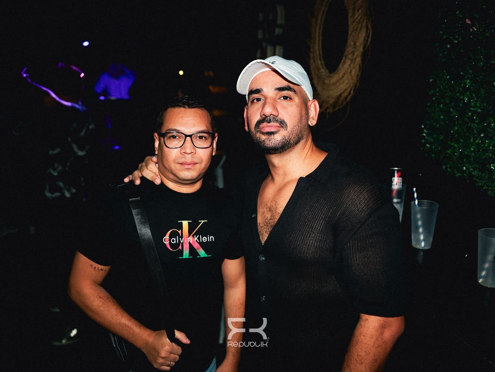 Dos personas posando juntas en un club con poca luz; una sostiene una bebida y lleva una gorra blanca.