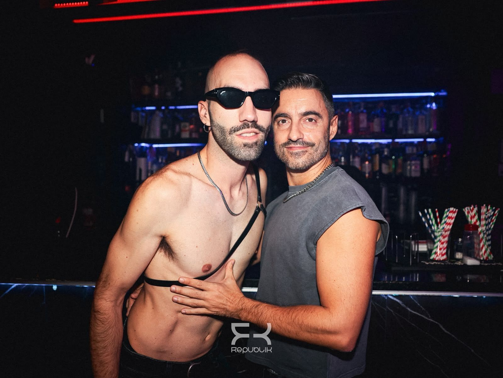 Dos hombres posan juntos en la barra de una discoteca con poca luz; uno va sin camisa y lleva gafas de sol.