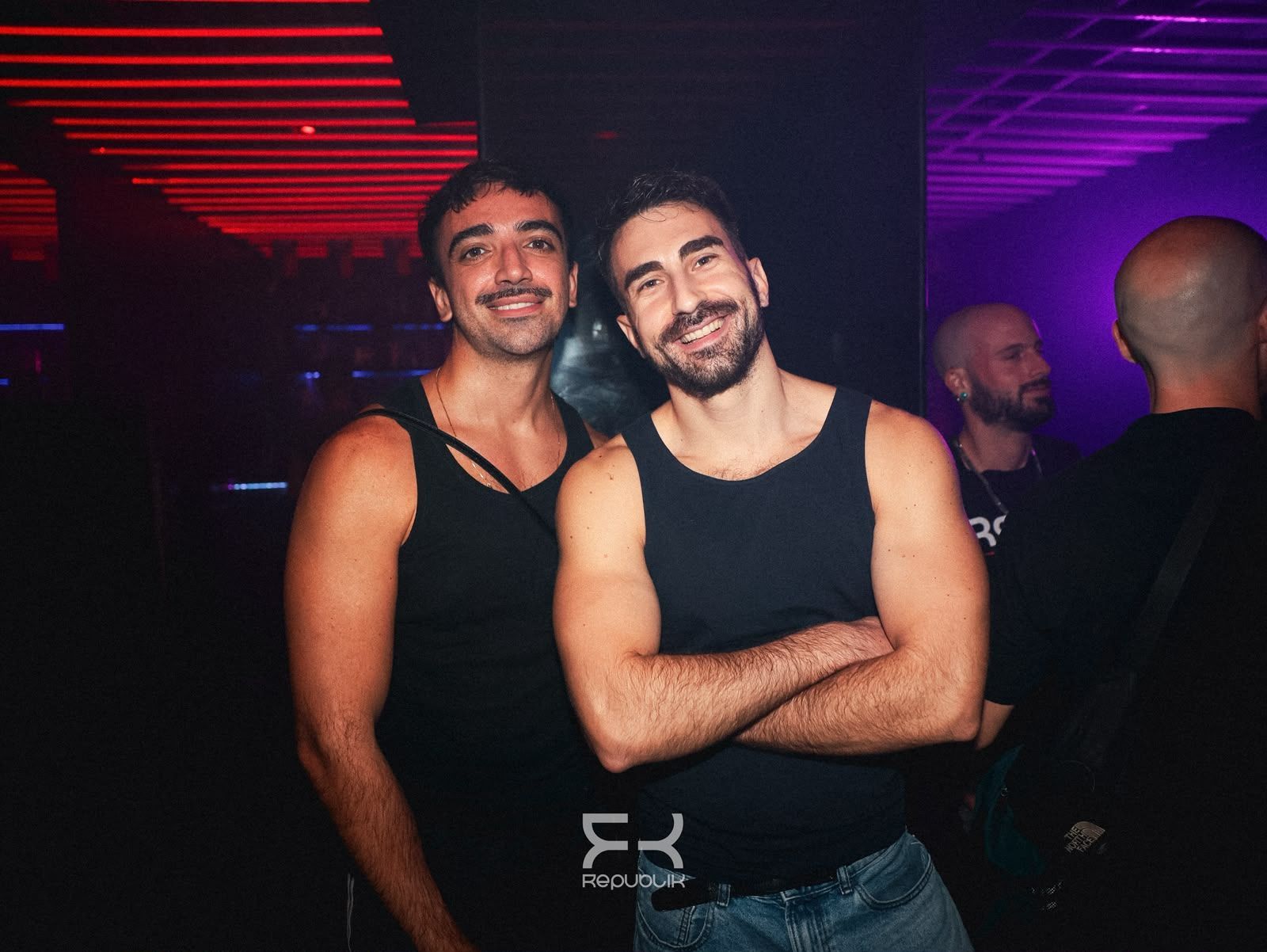 Dos hombres con camisetas negras sonríen y posan en una discoteca oscura con luces rojas y moradas