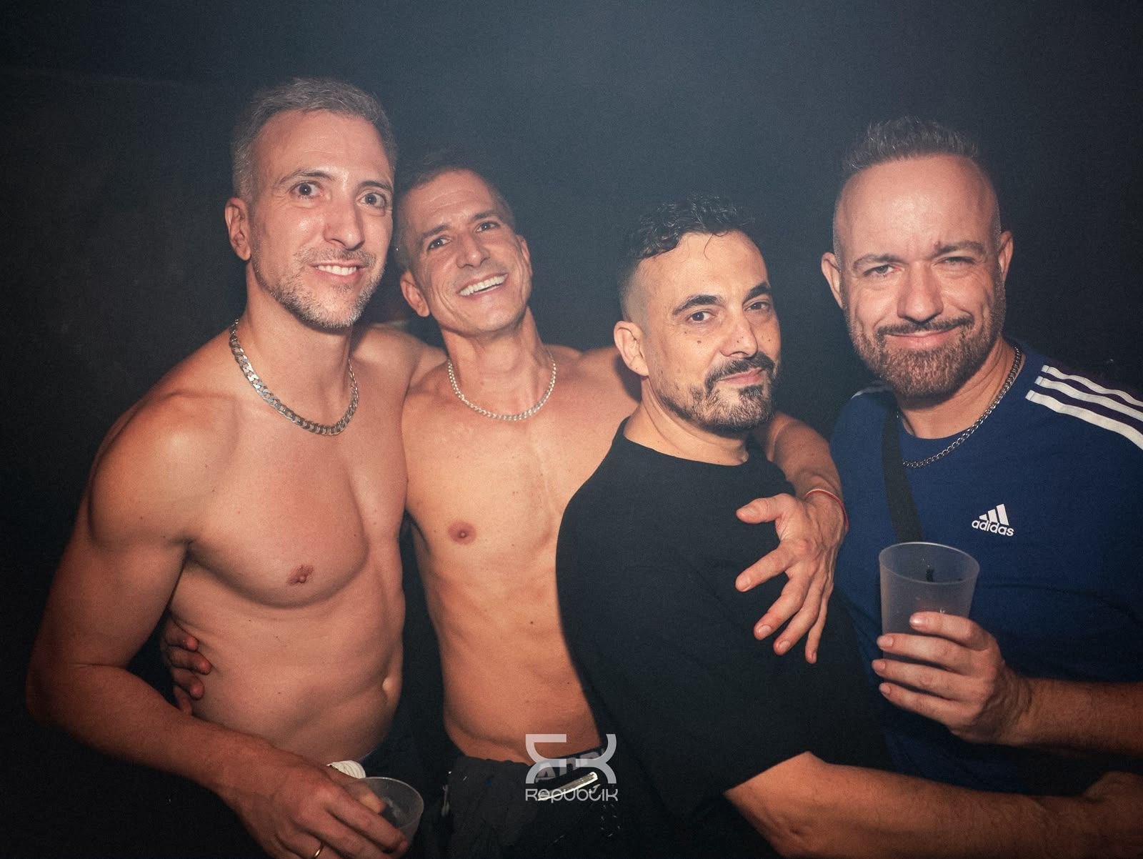 Cuatro hombres sin camiseta posando muy juntos y sonriendo en un ambiente con poca luz; uno sostiene una bebida