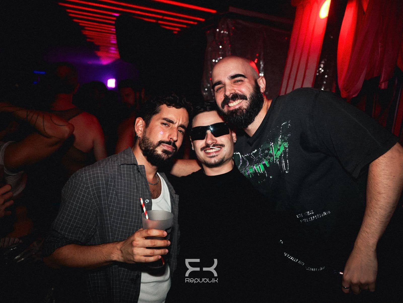 Tres personas sonríen en una fiesta con poca luz, bajo luces rojas y con bebidas en la mano.