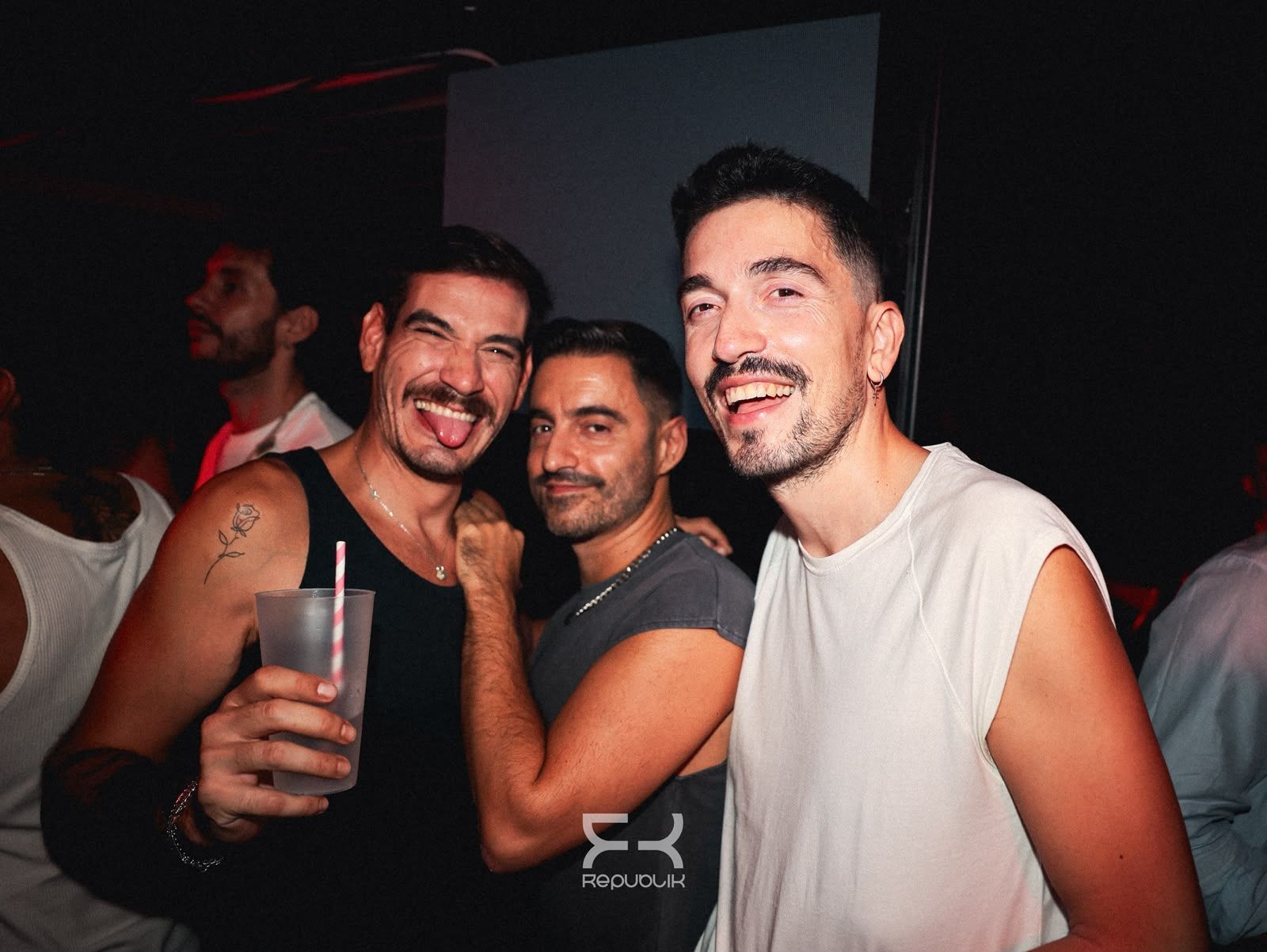 Tres personas sonríen y posan en una fiesta con poca luz; una de ellas sostiene una bebida.