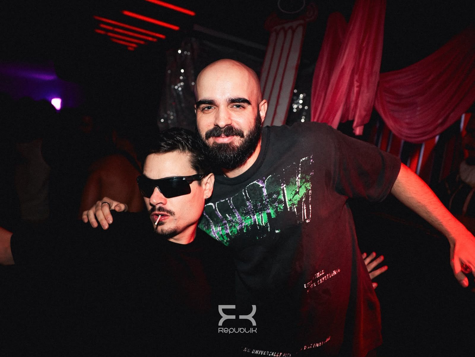 Dos hombres posando en un club con poca luz; uno lleva gafas de sol, con cortinas rojas e iluminación morada detrás de ellos