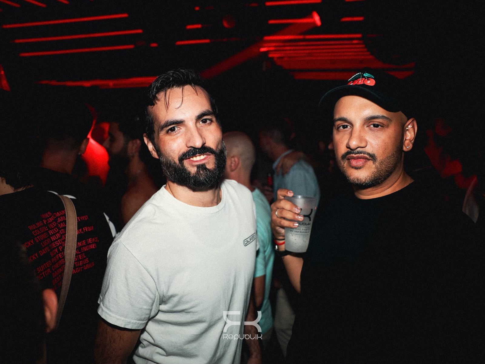 Dos hombres posando en una fiesta con poca luz roja, uno de ellos sosteniendo una bebida