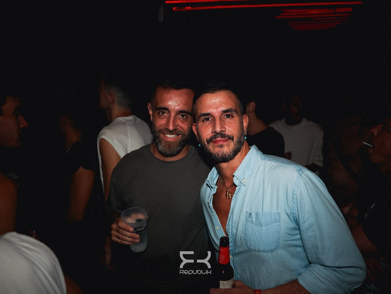 Dos hombres sonríen en una fiesta con poca luz; uno sostiene una copa y el otro, una botella, mientras unas luces rojas brillan sobre ellos.