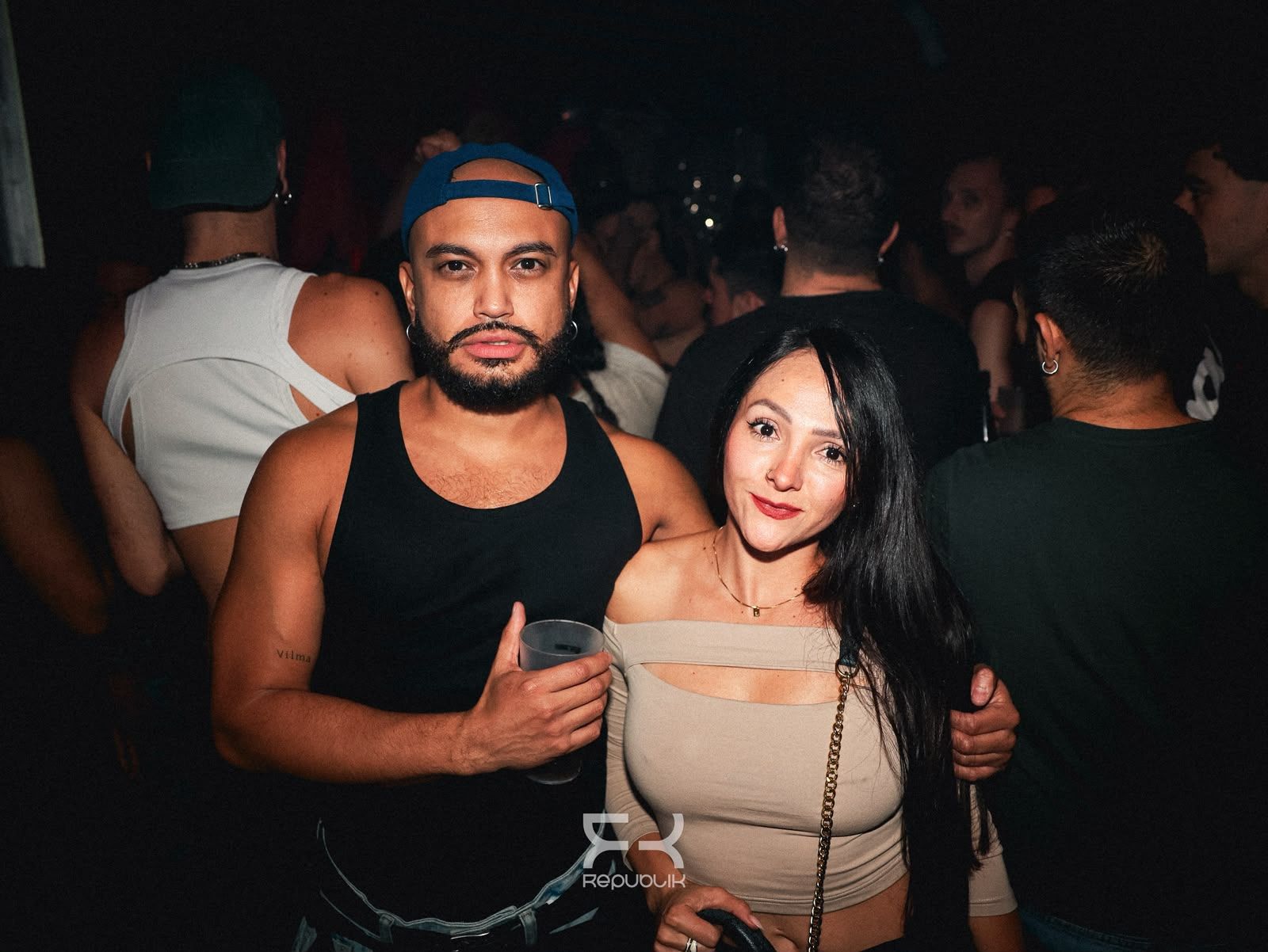 Dos personas posando en una discoteca con poca luz, con bebidas en la mano y mirando a la cámara.