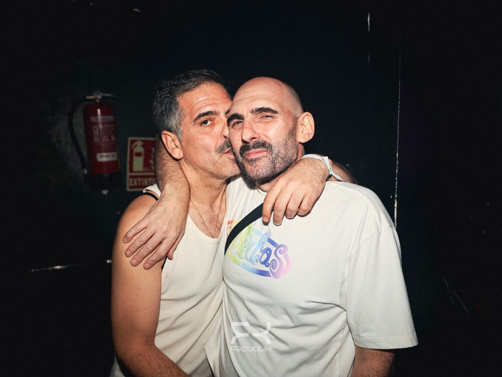 Dos personas abrazándose y sonriendo en una habitación con poca luz; una lleva una camiseta blanca y la otra, una camiseta sin mangas.
