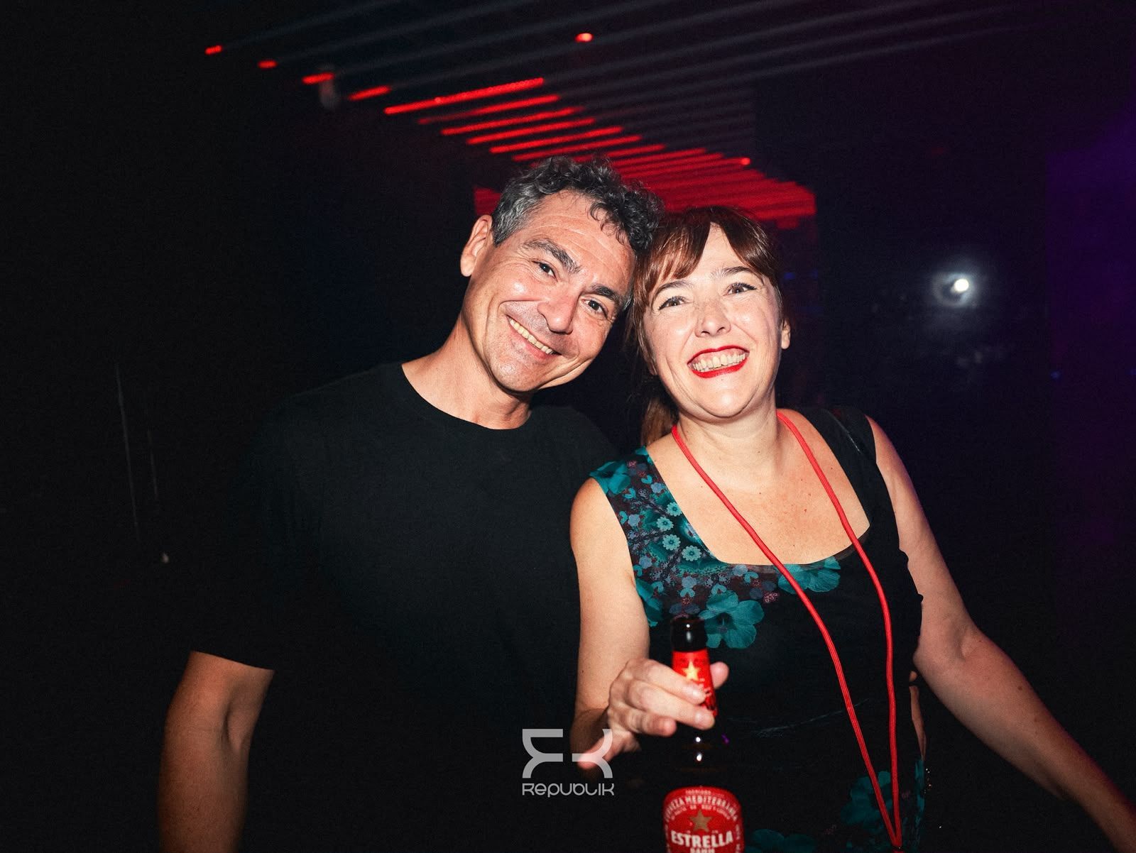 Dos personas sonríen en una discoteca oscura, con copas en la mano bajo luces rojas