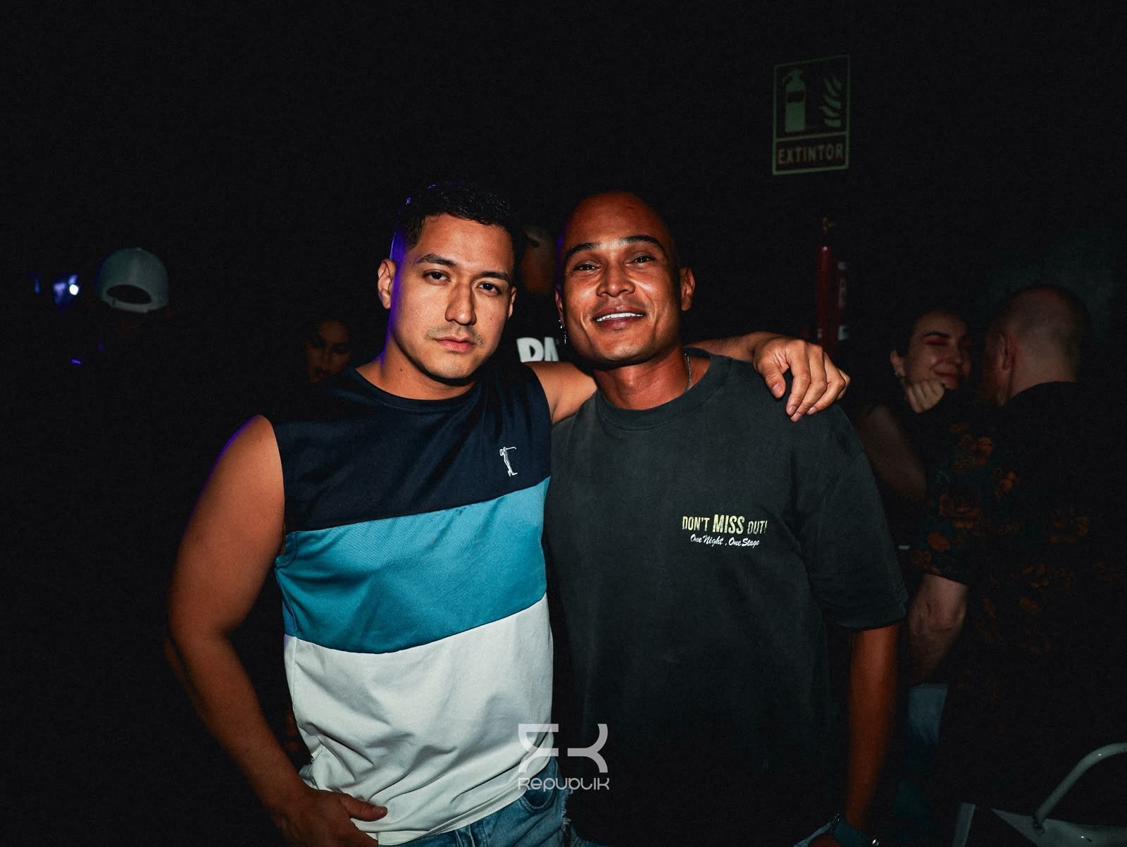 Dos amigos posando juntos en una discoteca poco iluminada; uno lleva una camisa azul y blanca y ambos sonríen.