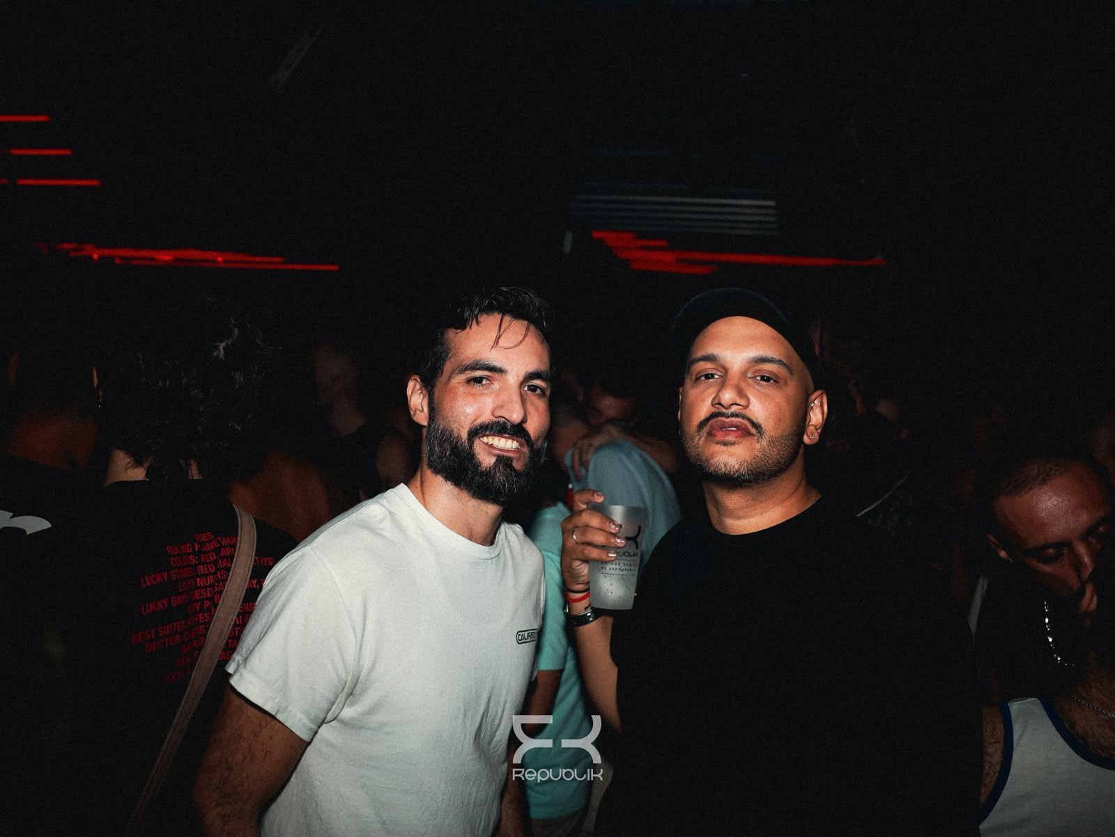 Dos hombres posando en una discoteca con poca luz, uno con camisa blanca y el otro de negro, con luces rojas de fondo