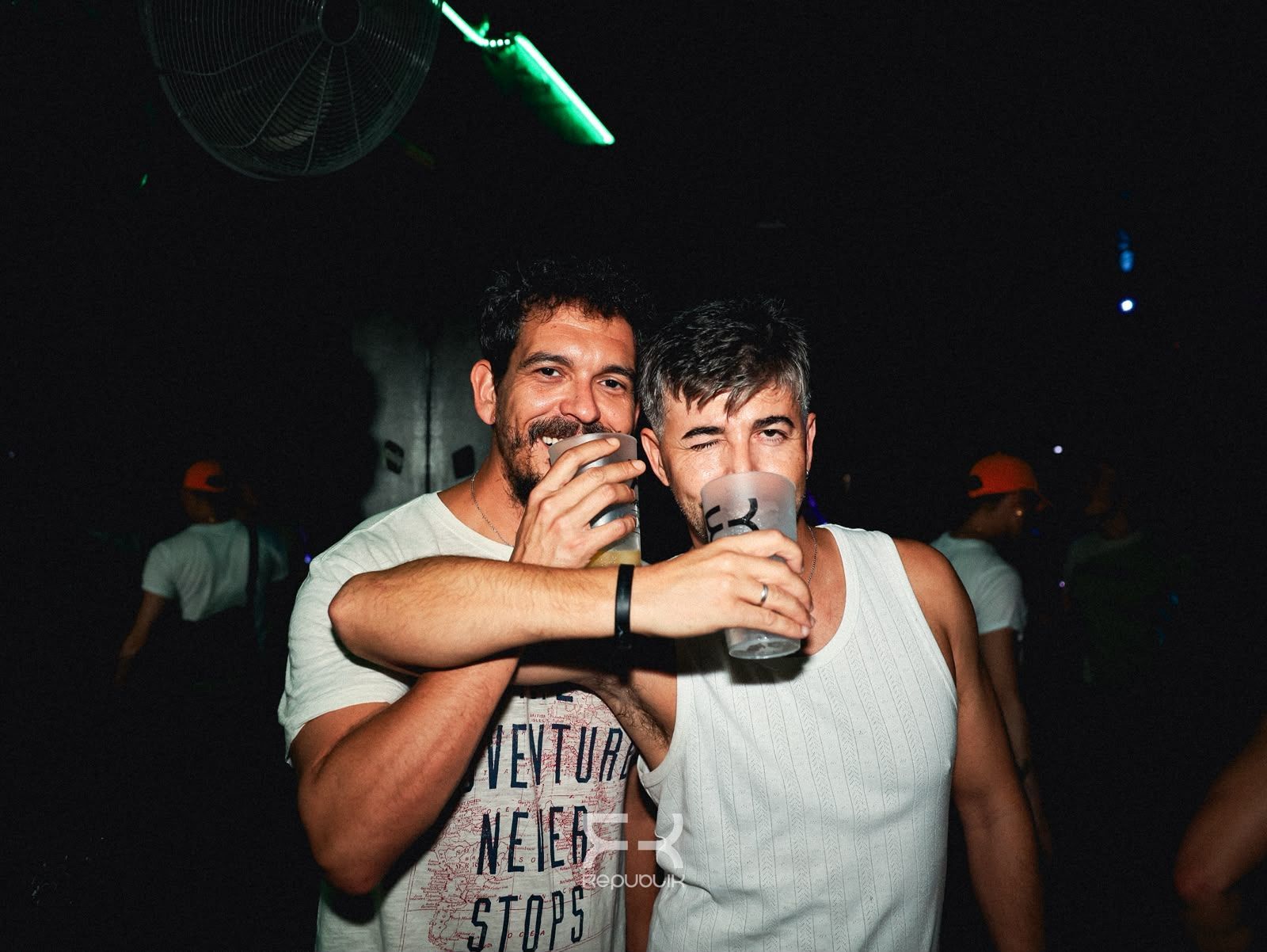 Dos hombres con camisas blancas brindan en una fiesta oscura con luces verdes.