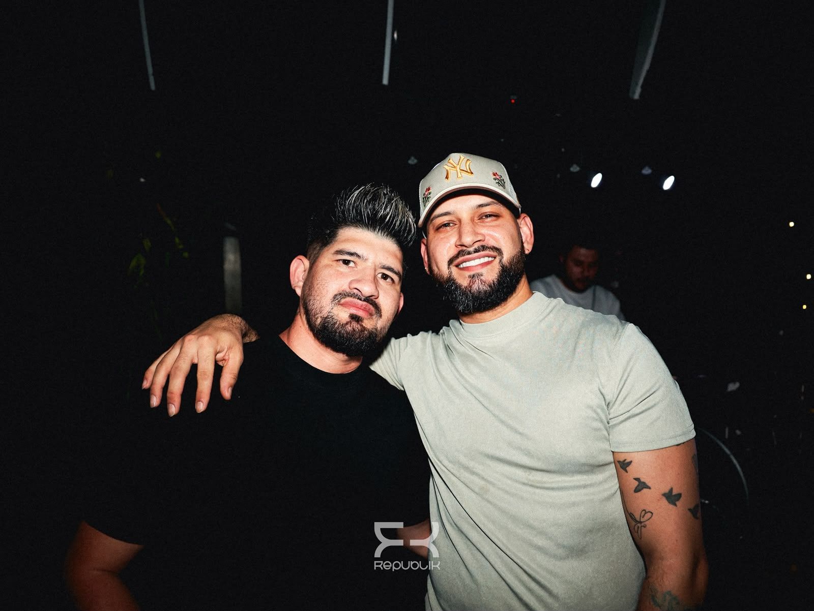 Dos amigos sonrientes posan juntos en un ambiente oscuro: uno lleva una camiseta negra y el otro, una camiseta clara y una gorra.