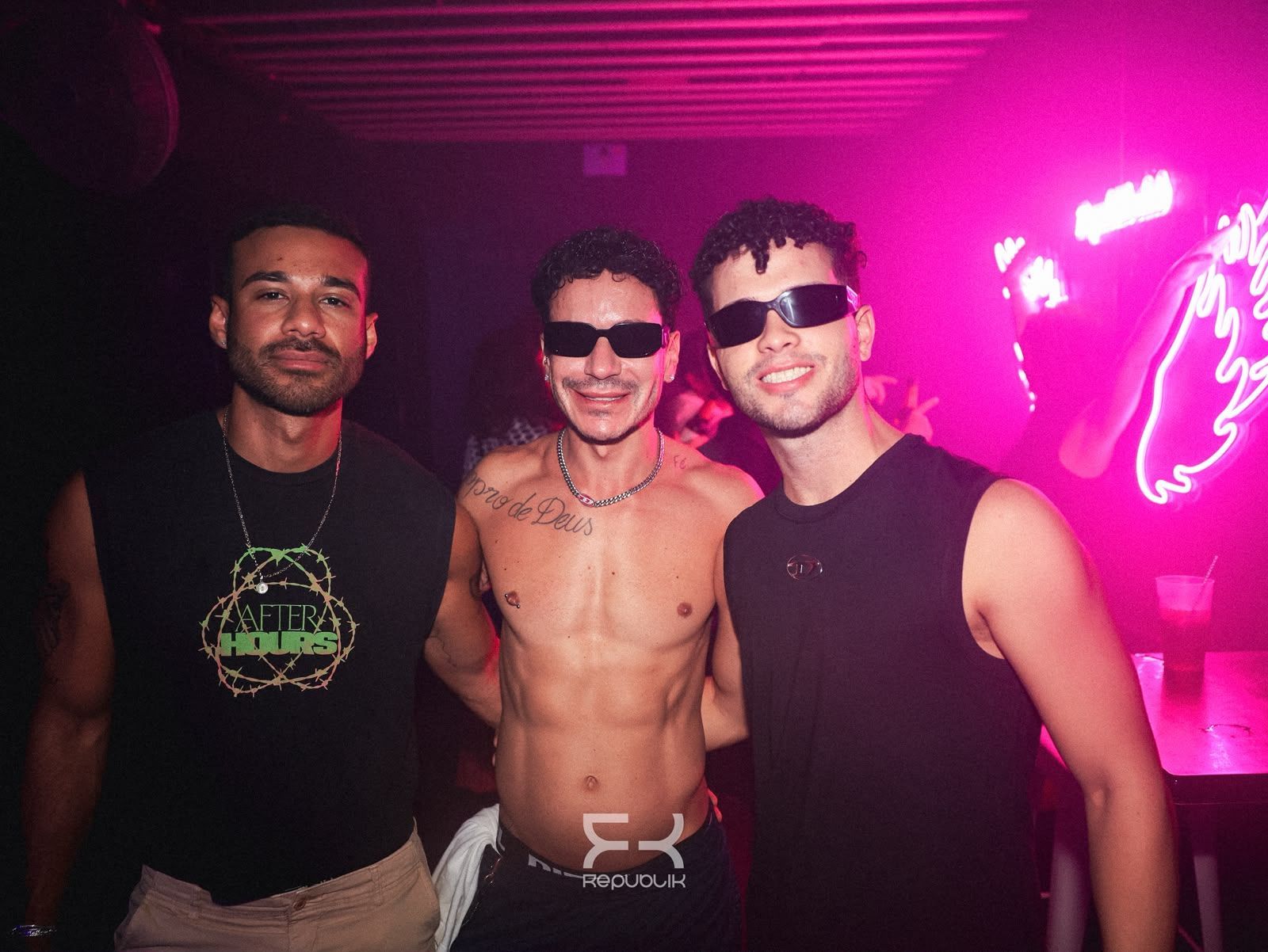 Tres hombres sin camiseta posan bajo la iluminación rosa neón de una discoteca; dos llevan gafas de sol.