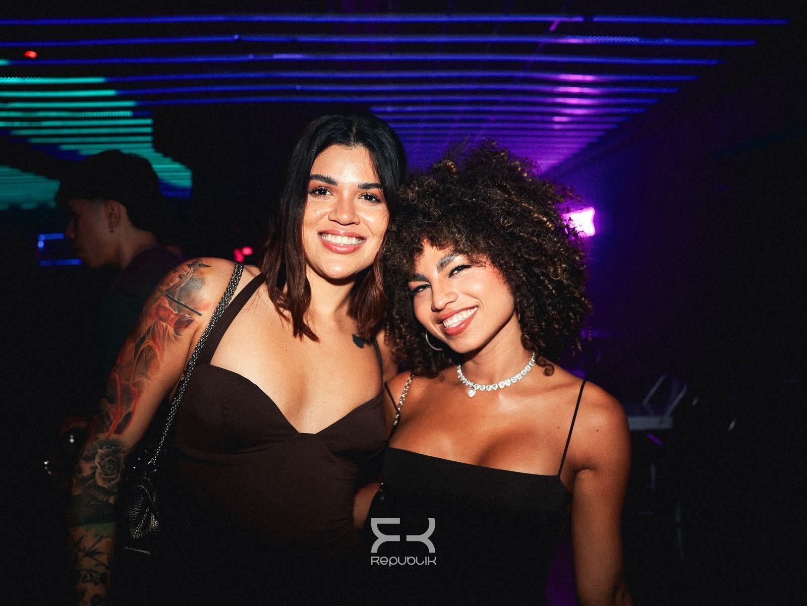 Dos personas sonríen en una discoteca a media luz, iluminada con luces moradas y luces de neón en el techo.