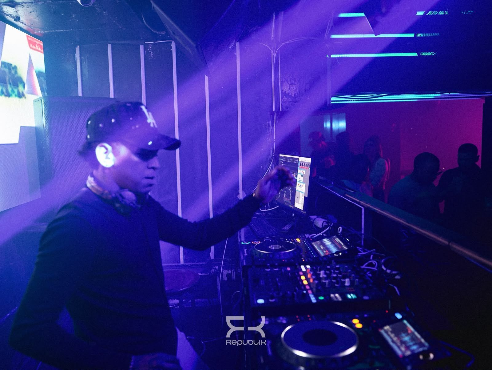 Un DJ actuando bajo luces moradas en una discoteca, con una mano levantada sobre una mesa de mezclas iluminada.