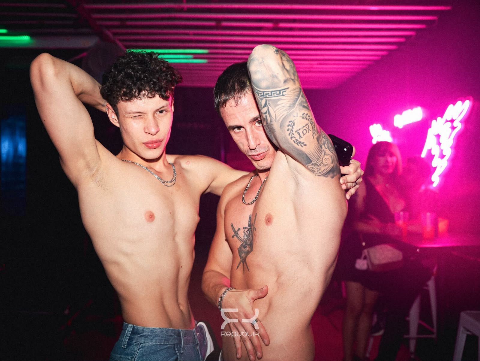 Dos personas sin camiseta posando en una discoteca iluminada con luces de neón rosas y un tatuaje visible en uno de los brazos