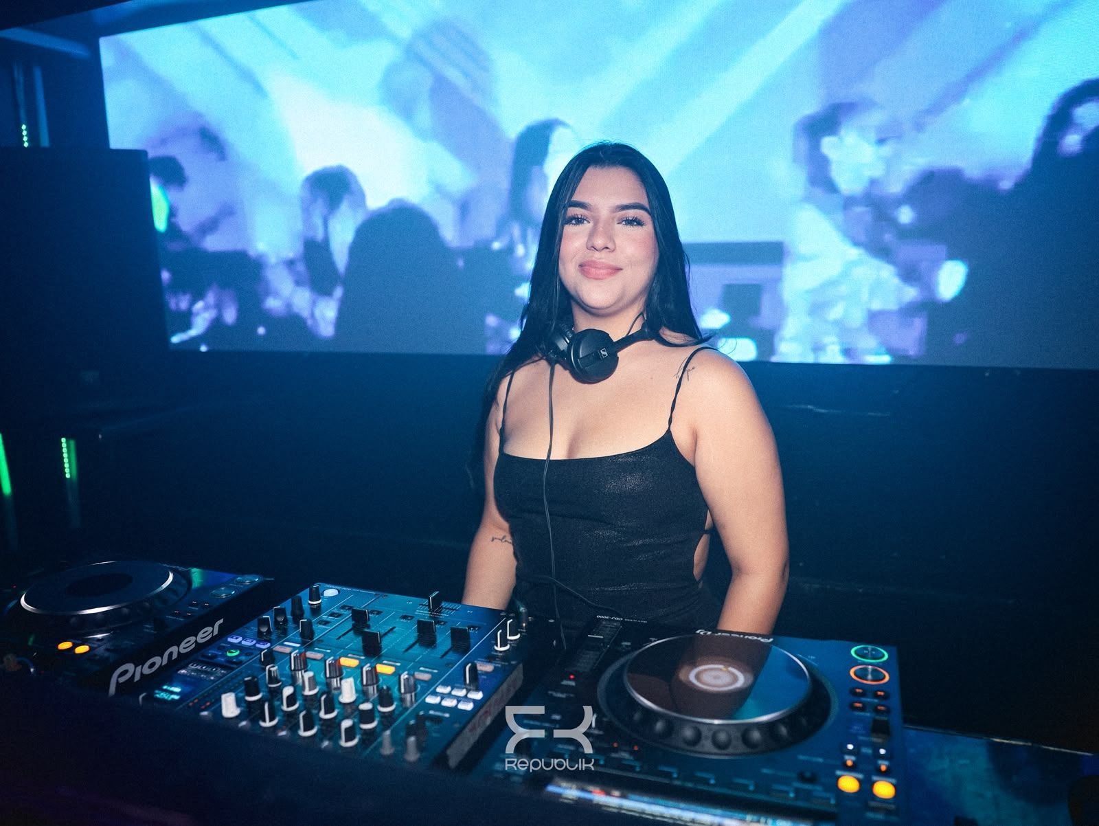 DJ en la cabina de una discoteca con auriculares, mesa de mezclas y luces azules de escenario al fondo