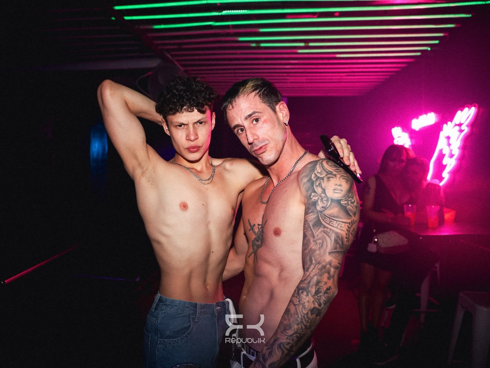 Dos personas sin camisa posando en una discoteca iluminada con luces de neón rosas y verdes