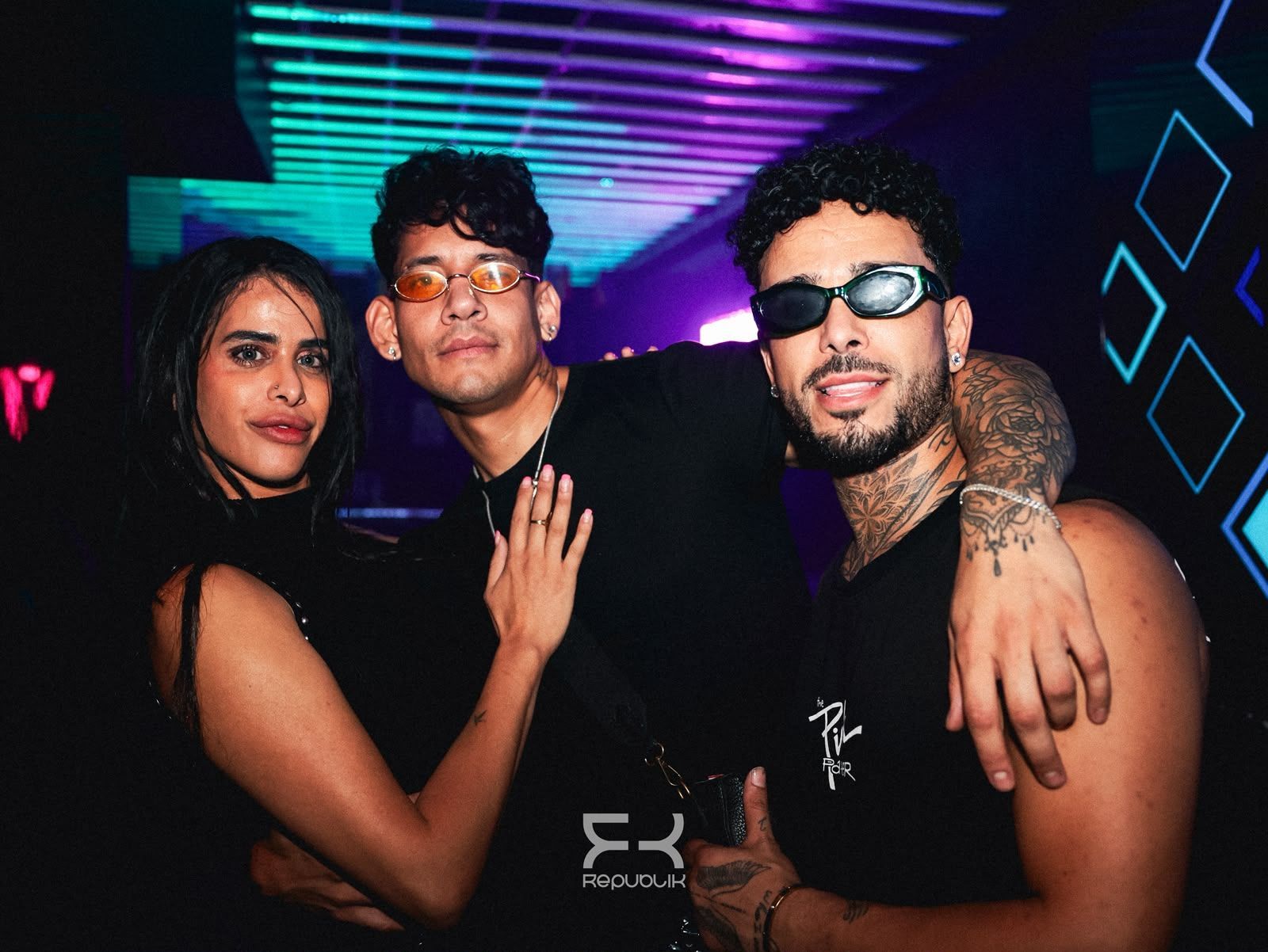 Tres personas posando en una discoteca iluminada con luces de neón azules y moradas al fondo.
