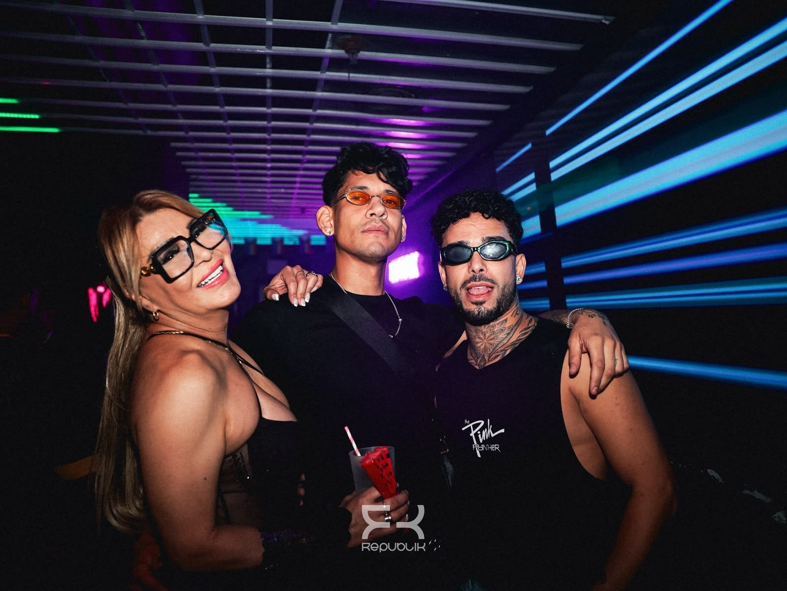 Tres amigos posando en un túnel iluminado con luces de neón, sonriendo y con gafas de sol.