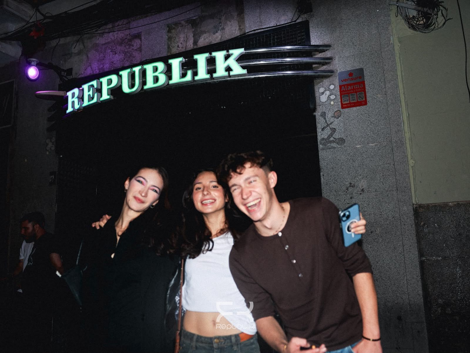 Tres amigos sonríen y posan frente a la entrada de un club, bajo un letrero que dice “REPUBLIK”