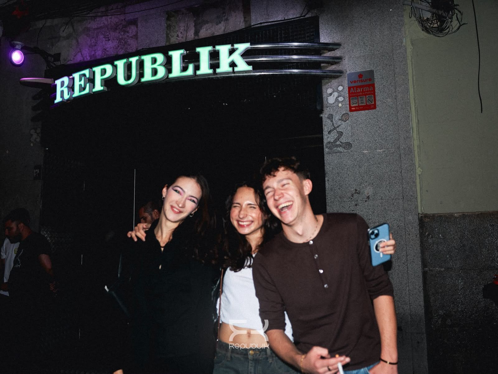 Tres amigos sonríen frente a un local con un letrero luminoso que dice “REPUBLIK” por la noche