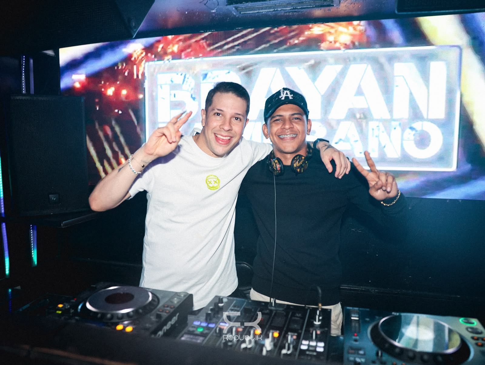 Dos DJ sonrientes posando detrás de una mesa de mezclas en una cabina de club iluminada con luces de neón