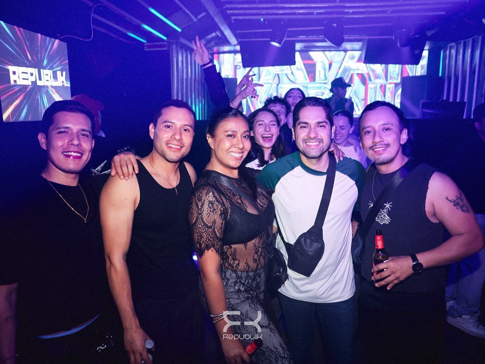 Grupo de seis amigos sonrientes posando en una discoteca iluminada con luces de neón moradas y azules.