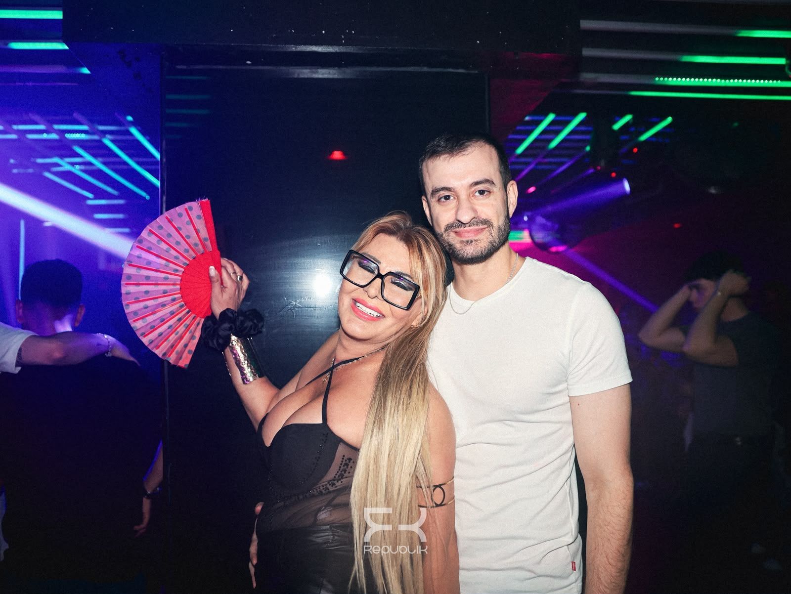 Dos personas sonríen en una discoteca iluminada con luces de neón; una sostiene un abanico rojo.