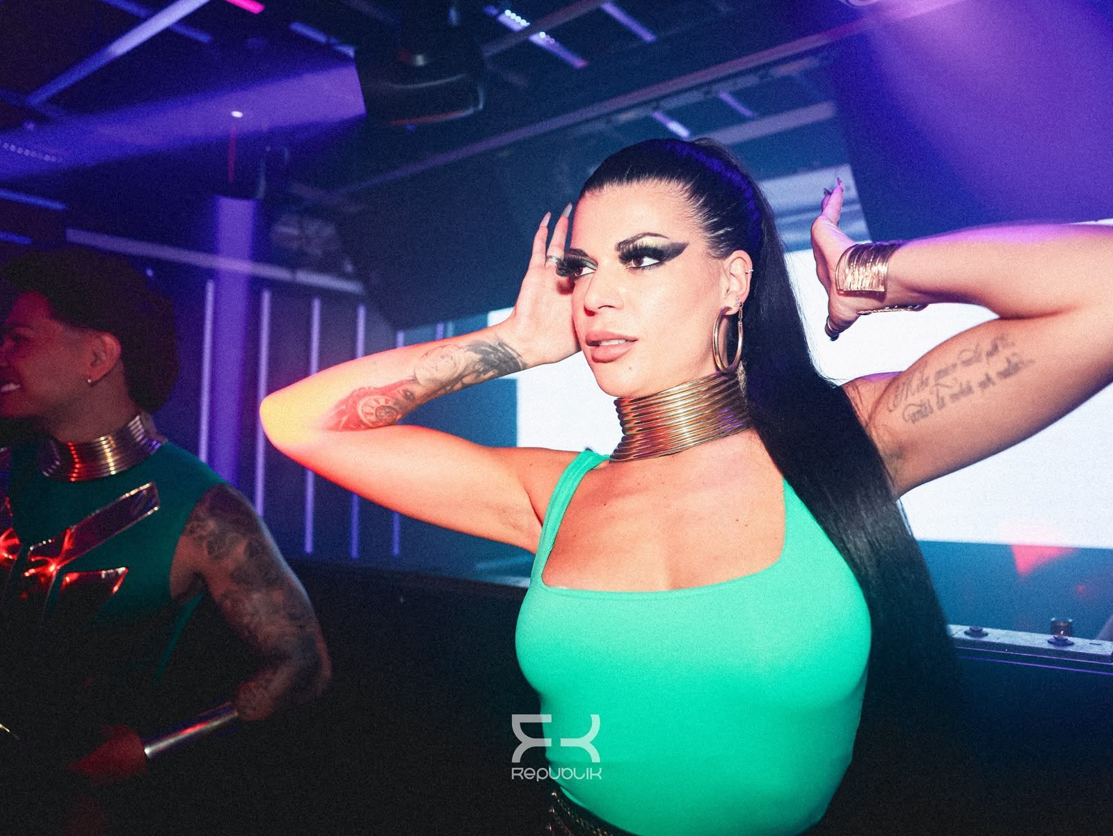Mujer con una blusa verde neón posando con las manos detrás de la cabeza en un club con poca luz y luces moradas