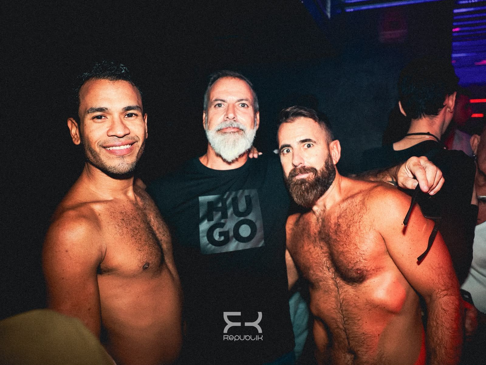 Tres hombres sin camisa posan juntos en un gimnasio o club con poca iluminación; uno de ellos lleva una camiseta negra y está en el centro.
