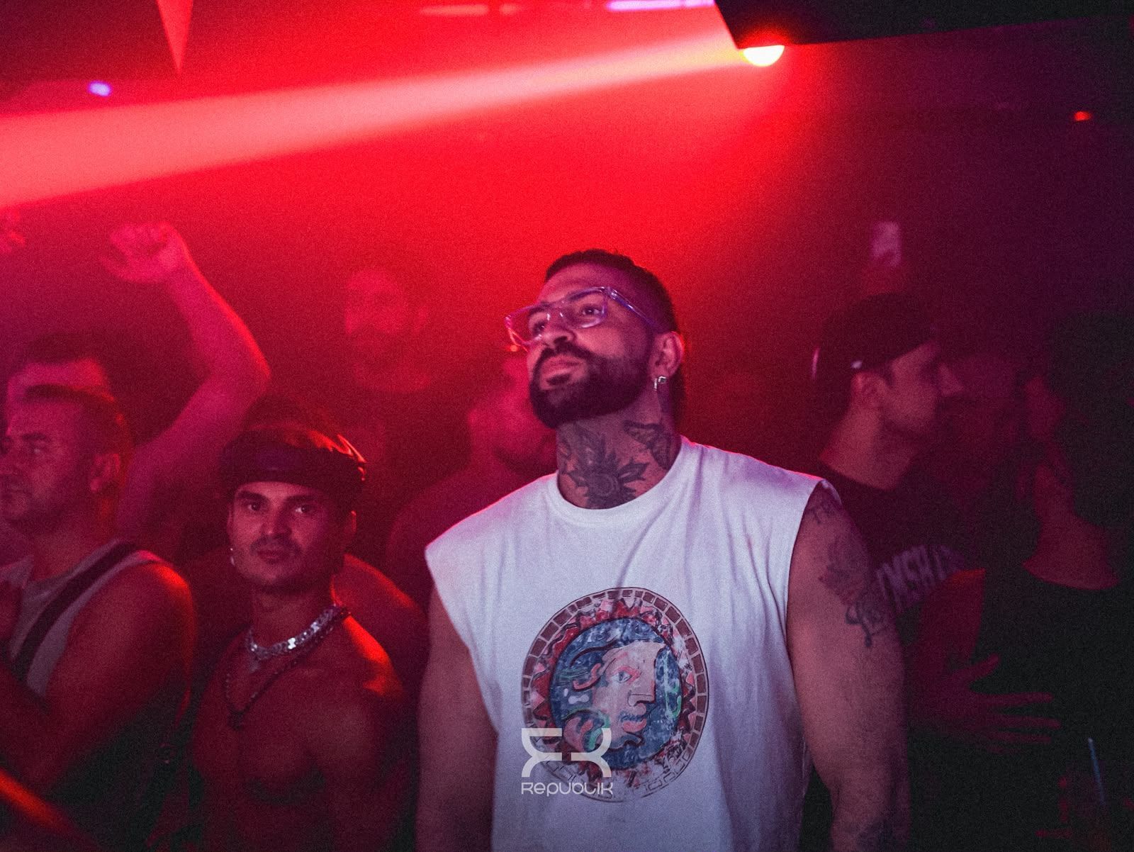 Multitud en una discoteca iluminada de rojo, con un hombre en una camiseta blanca sin mangas en el centro mirando hacia arriba