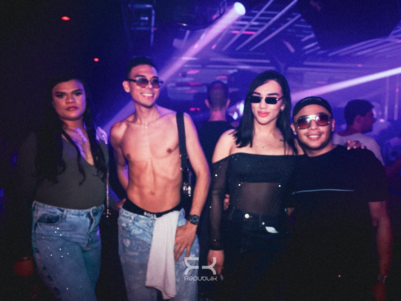 Cuatro personas posando en una discoteca iluminada de color morado; dos llevan gafas de sol y, delante, hay un hombre sin camiseta.