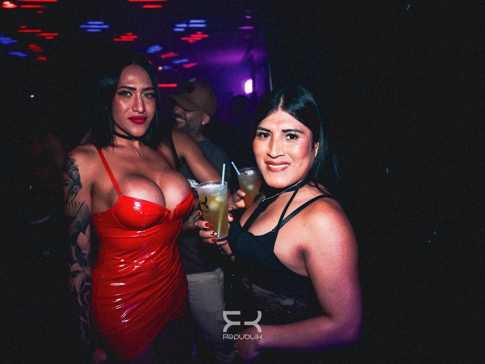 Dos mujeres en una discoteca, una con un vestido rojo y otra de negro, con bebidas en la mano bajo luces de colores