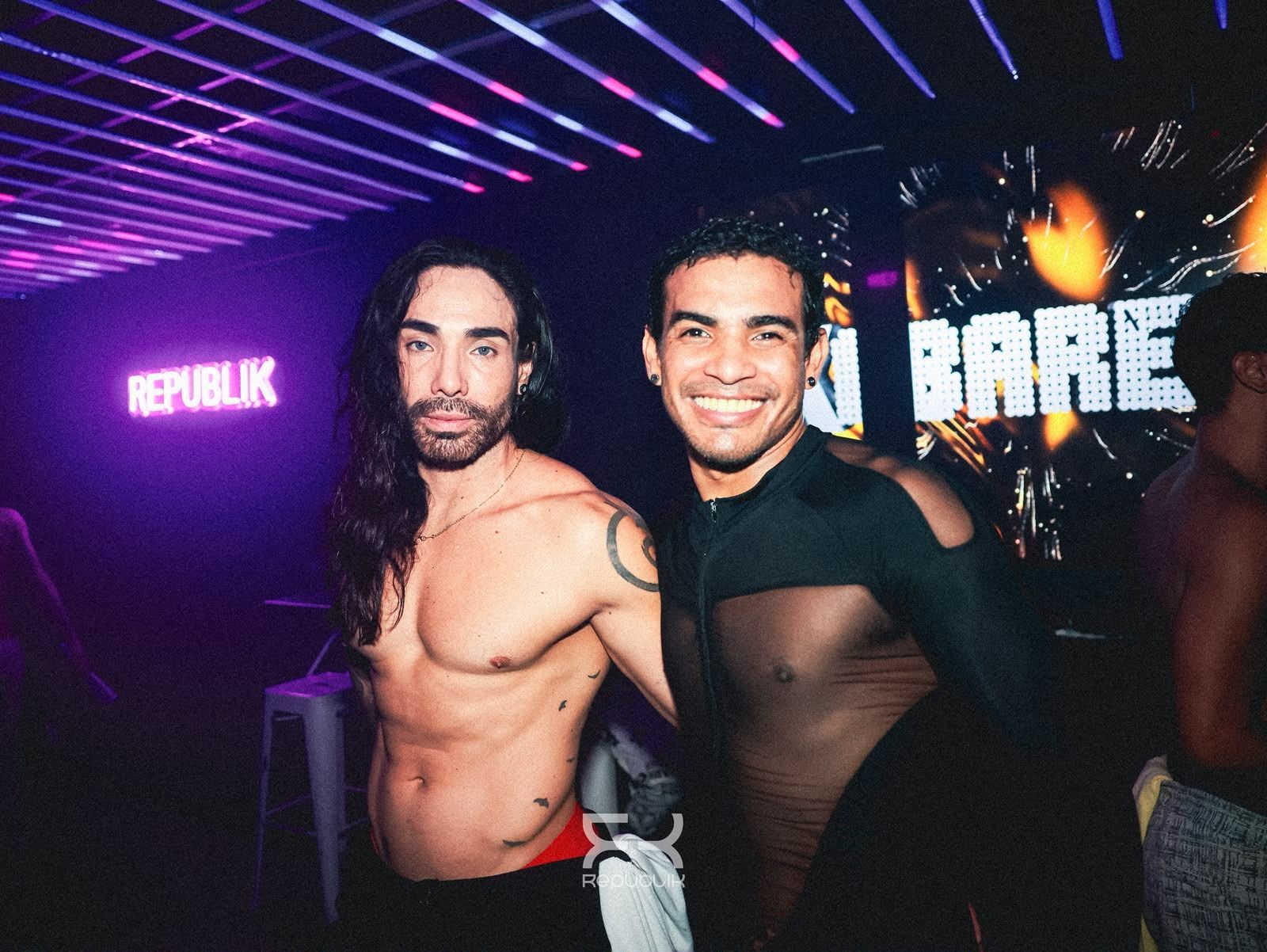 Dos personas posando en una discoteca iluminada con luces de neón, una sin camiseta y la otra con una blusa negra transparente, sonriendo.