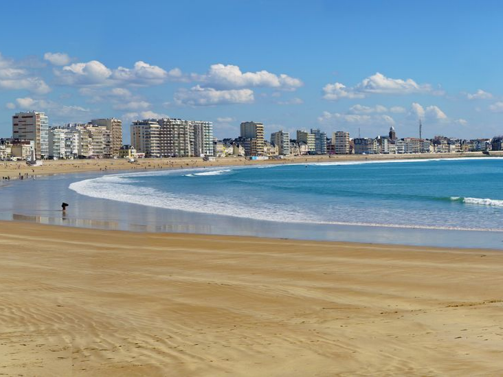 Plage ensoleillée au sable doré, avec la ville en fond sous un ciel partiellement nuageux