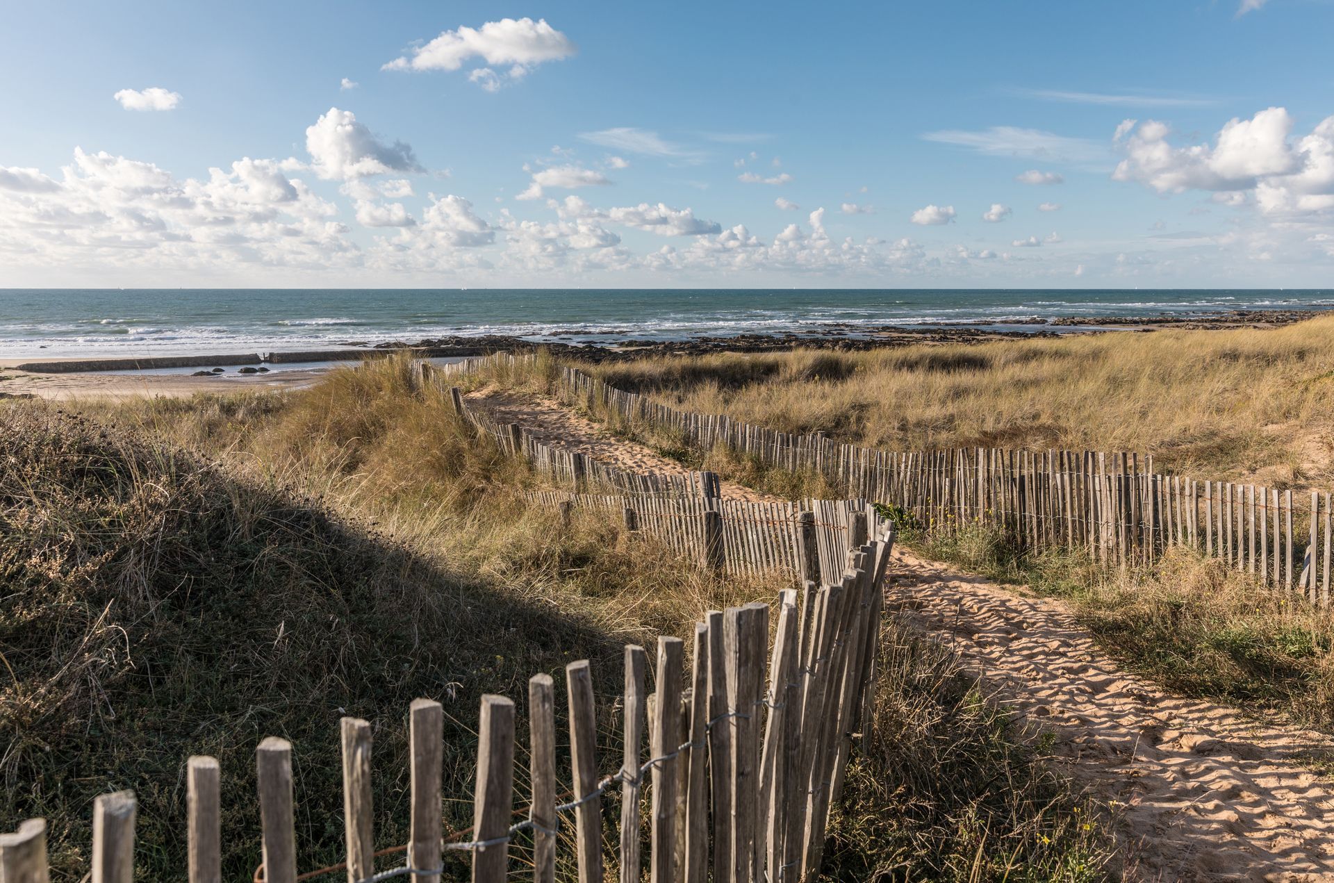 Sentier sablonneux traversant des dunes bordées de clôtures en bois, menant à un à la plage sous un ciel bleu.