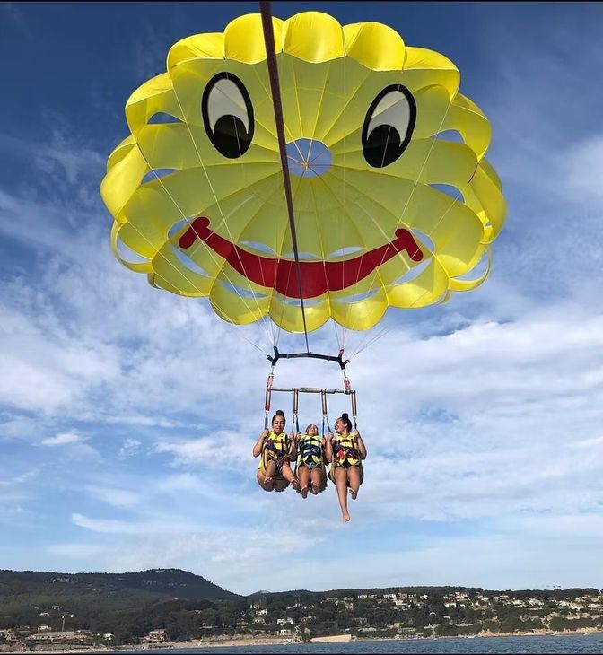 Un parachute jaune imprimé smiley transporte quatre personnes au-dessus de la mer près d'une ville côtière.