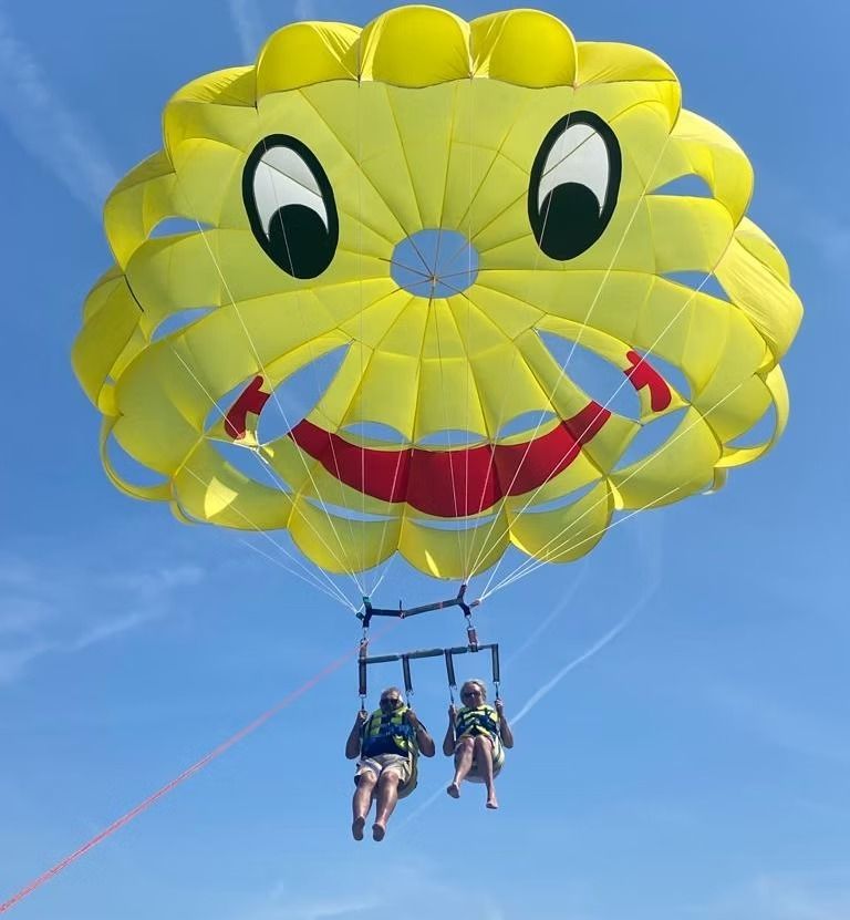 Deux personnes font du parachute ascensionnel sous une grande toile jaune en forme de smiley sur fond de ciel bleu.