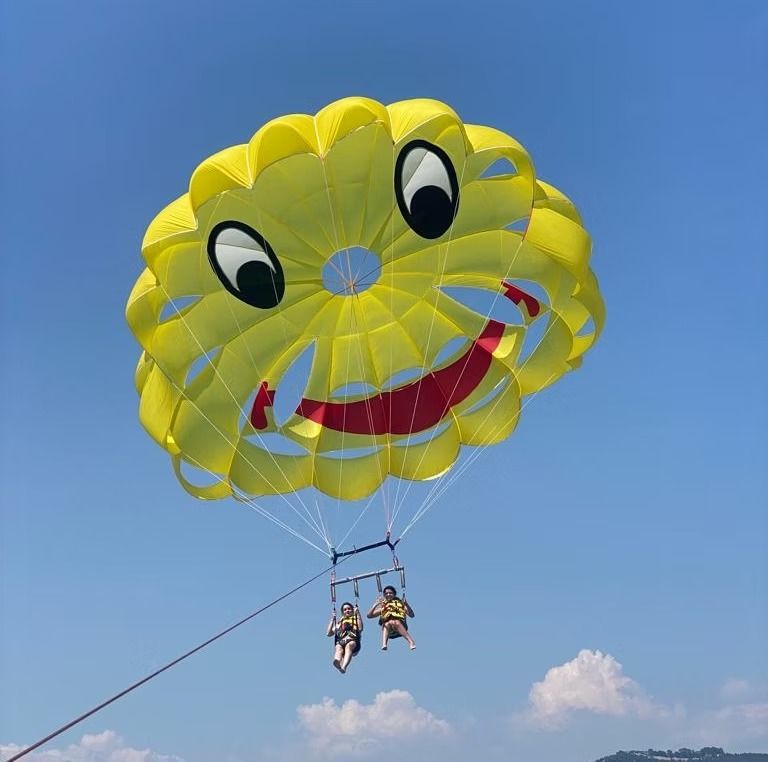 Deux personnes font du parachute ascensionnel sous une grande toile jaune en forme de smiley sur fond de ciel bleu.