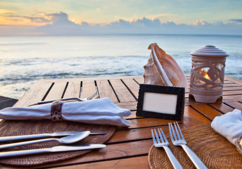 Table set for a seaside dinner with a seashell, candle, and ocean view at sunset.