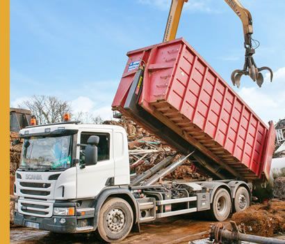 Un camion blanc avec une benne rouge est chargé de ferraille par une excavatrice.