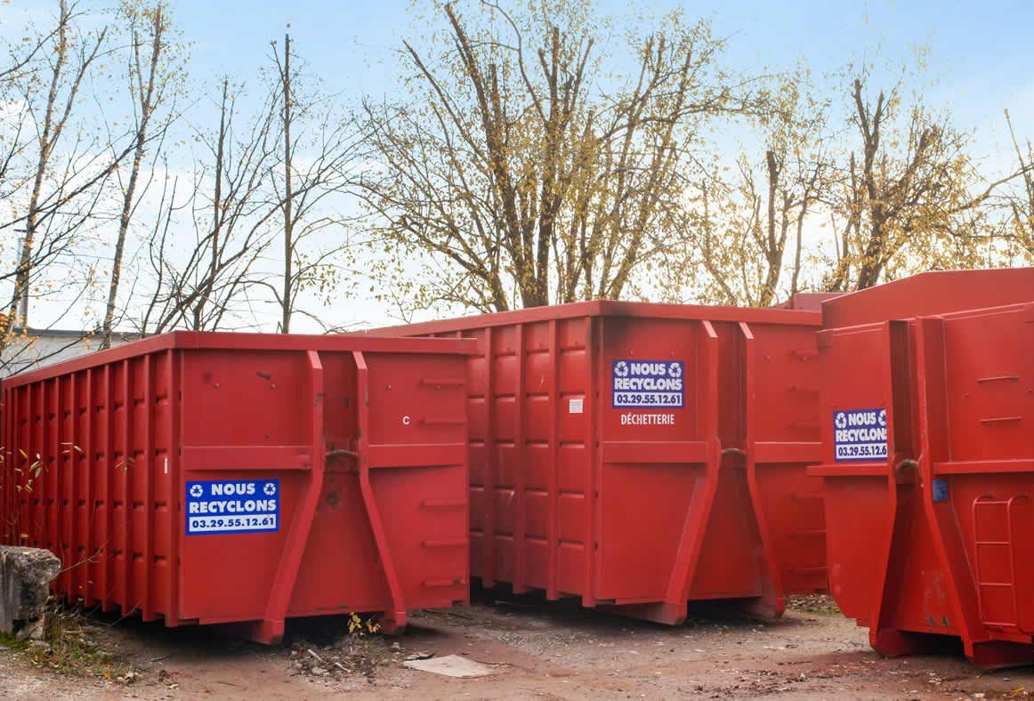 Des bennes à ordures industrielles rouges se trouvent à l'extérieur, probablement dans une installation de gestion des déchets, avec des arbres en arrière-plan.