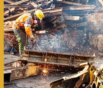 Un ouvrier portant un gilet de sécurité et un casque de chantier découpe du métal dans une casse, des étincelles jaillissent.