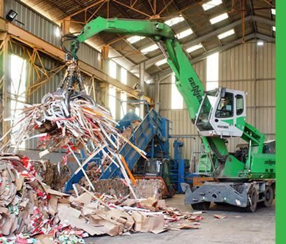 Une pelle mécanique industrielle verte soulève un tas de carton et de papier dans une installation de recyclage.