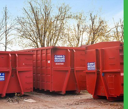 Des conteneurs de recyclage rouges à l'extérieur, avec des arbres sans feuilles en arrière-plan.
