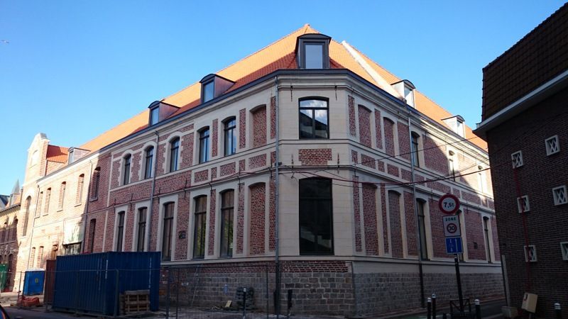 Bâtiment historique en briques, orné de fenêtres et d'un toit pentu, situé à un angle de rue ensoleillé.