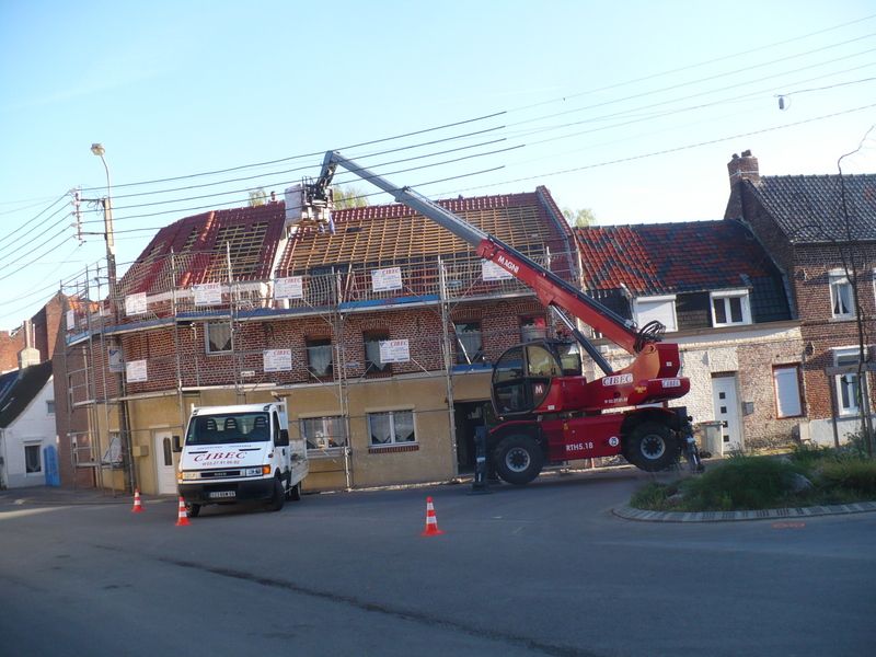 Un camion-grue rouge soulève des débris à côté d'une maison en briques après des dégâts au niveau du toit ; une camionnette de chantier et des cônes de signalisation se trouvent à proximité.